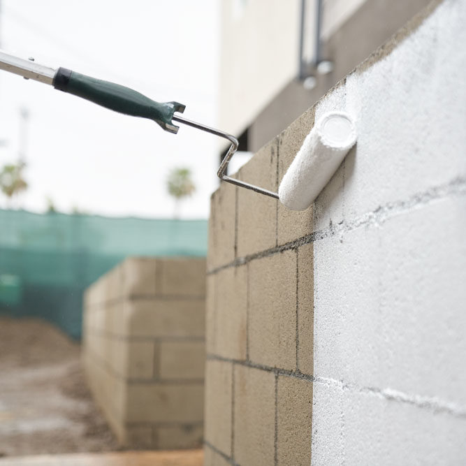 Image of paint roller applying primer to cinderblock wall outside.